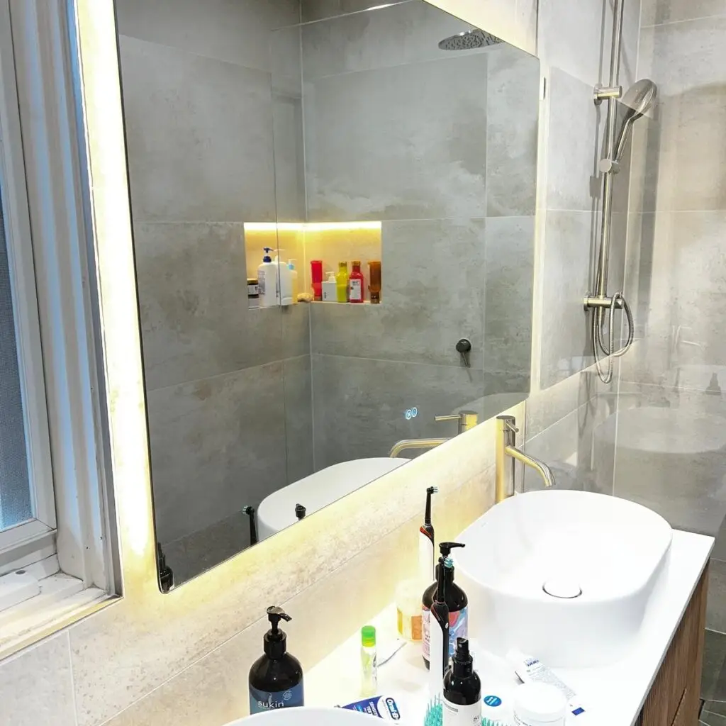 Modern bathroom vanity with backlit mirror and white vessel sink, cluttered countertop toiletries nearby.